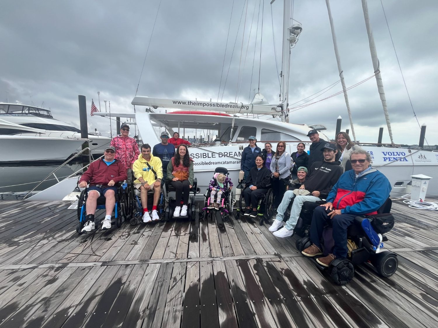 Picture of a Group of wheelchair users and non-wheelchair users in front of a docked white catamaran on a cloudy day.