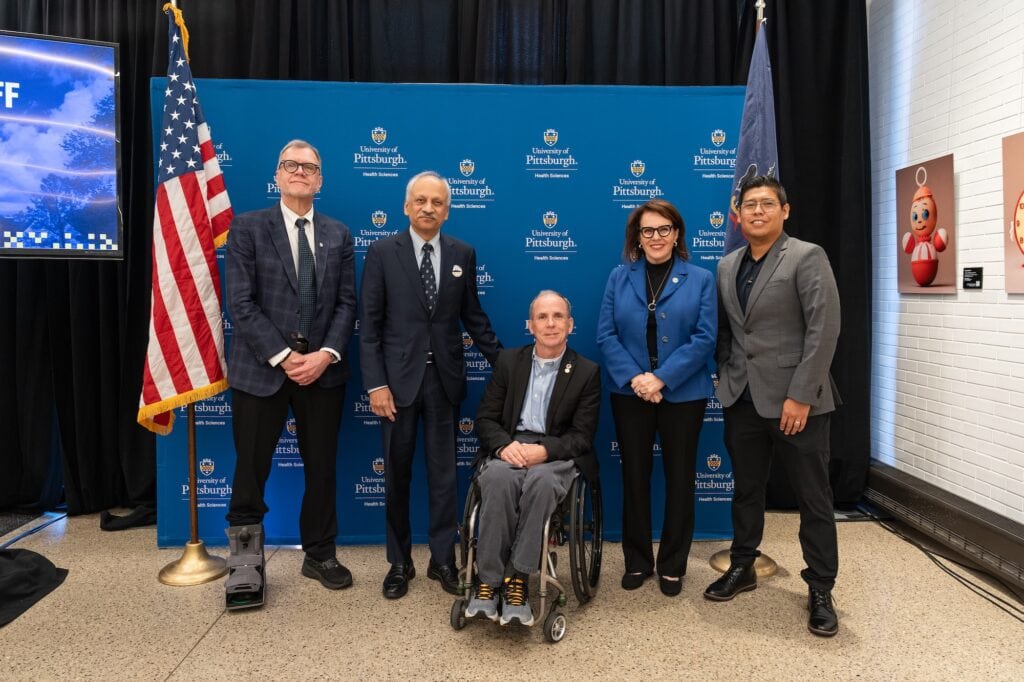 ID: a group of people posing for the photo. A wheelchair user is in the middle. A background of an American flag a black curtain and a blue wall they're all standing in front of.