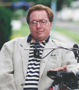 Image description: this is a photograph of Girard in his wheelchair wearing a White coat and stripe shirt.