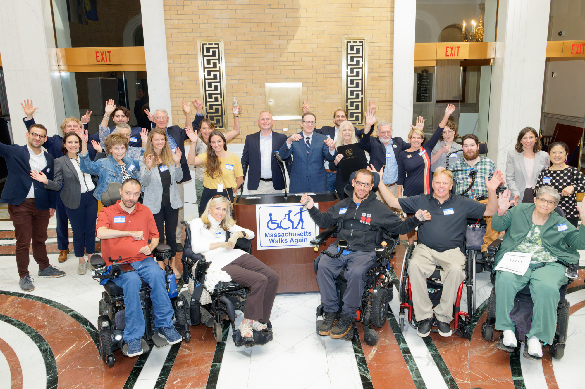 Image description: a group of wheelchair users and non-wheelchair users gathered around a podium at the Massachusetts hall of flags in the state house.